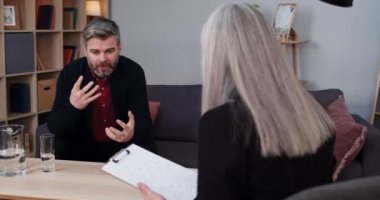 Olgun bir adamın kanepede oturup bir kadınla terapi seansı yapması. Kadın psikolog notlar alırken, erkek sakallı insan duygusal ve hareketli konuşuyor..