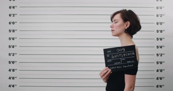 Mugshot de mujer joven criminal con corte de pelo corto girando la cabeza y mirando a la cámara mientras está de pie a un lado. Recorte vista de morena pesón femenino sosteniendo cartel para la foto en el departamento de policía. — Vídeos de Stock