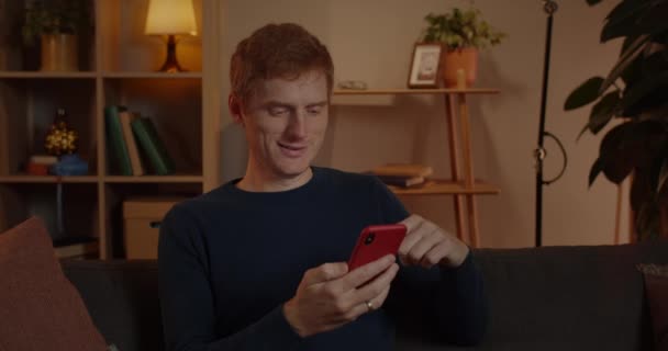 Vista de la cosecha de hombre de pelo rojo positivo usando y desplazando la pantalla del teléfono inteligente mientras está sentado en el sofá en casa. Guapo chico sonriendo y cheking contenido del teléfono mientras pasa tiempo libre. — Vídeos de Stock