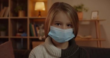 Kameraya bakan tıbbi koruyucu maskeli genç bir kızın portresi. Evde oturan uzun sarı saçlı güzel bir çocuğun yakın görüntüsü. Karantina ve virüs kavramı.