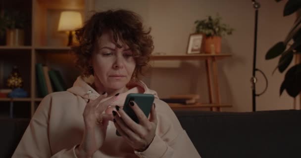 Crop view of excited woman with short curly hair scrolling phone screen while sitting on sofa at home. Vrolijke vrouwelijke persoon verheugen zich tijdens het gebruik en het controleren van smartphone. — Stockvideo