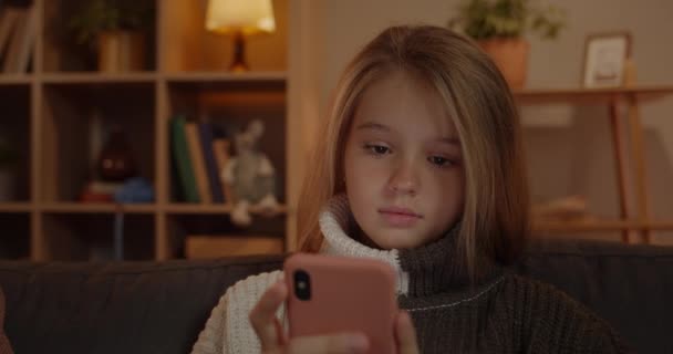 Nært syn på unge søte jenter som ser på smarttelefon mens de sitter på sofaen hjemme. Vakkert barn med langt blondt hår bruker telefon mens de tilbringer fritiden . – stockvideo