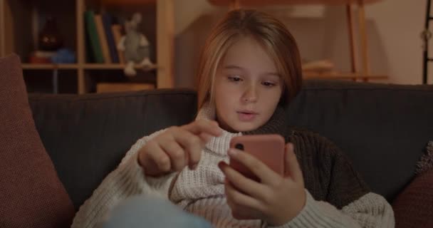 Крупним планом вид на здивовану маленьку дитину, що прокручує екран телефону і каже "Ого", сидячи на дивані вдома. Досить молода дівчина з довгим світлим волоссям використовує смартфон і виглядає здивованим . — стокове відео