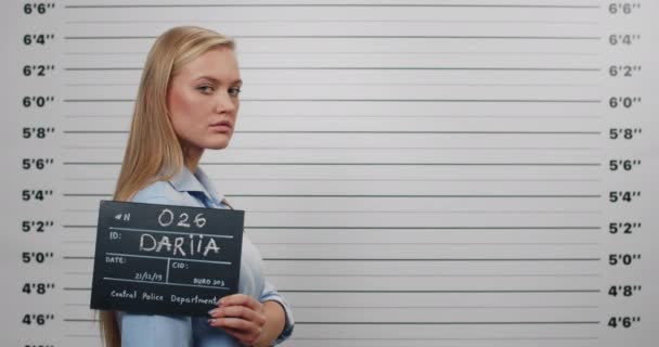 Side profile mugshot of millennial woman turning head and looking to camera. Female arrested holding sign for photo while standing in front of police metric lineup wall. — Stock Video