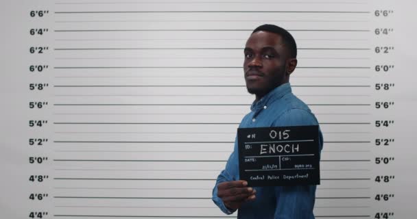 Ληστεία ενός Αφροαμερικανού που γυρνά στο πλάι ενώ κρατάει ταμπέλα και ποζάρει για φωτογραφία. Crop άποψη της εγκληματικής αρσενικό πρόσωπο με γενειάδα κοιτάζοντας την κάμερα στο αστυνομικό τμήμα. Έννοια του εγκλήματος. — Αρχείο Βίντεο