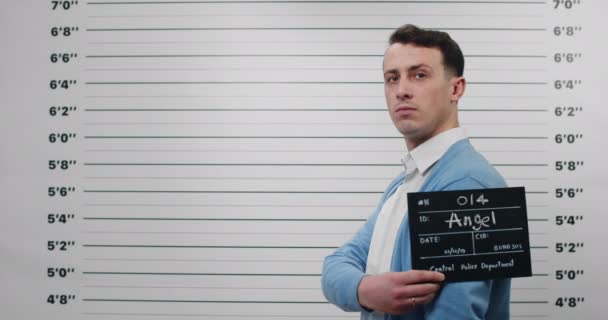 Mugshot de la persona masculina en camisa y cárdigan girando la cabeza mientras está de pie a un lado mientras sostiene el cartel y posando para la foto. Detenido hombre con el pelo corto ondulado mirando a la cámara en la estación de policía. — Vídeo de stock