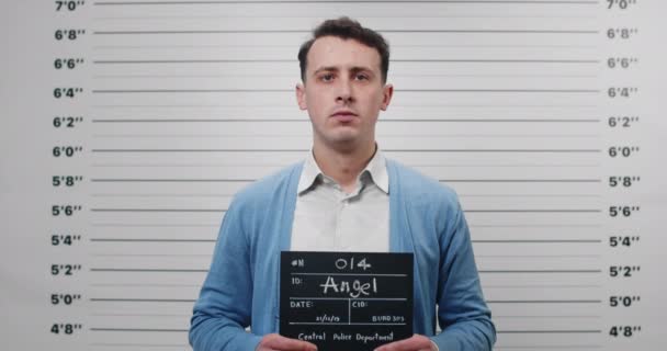 Retrato del hombre con camisa y cárdigan sosteniendo el cartel de la foto en el departamento de policía. Vista de la cosecha de la persona masculina en los años 30 posando, levantando la cabeza y mirando a la cámara. Concepto de mugshot y crimen. — Vídeos de Stock