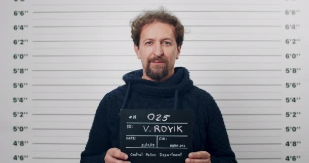 Retrato del hombre barbudo adulto sosteniendo letrero para la foto en el departamento de policía. Vista de la cosecha de la persona masculina con ojos azules posando, levantando la cabeza y mirando a la cámara. Concepto de mugshot y crimen. — Vídeos de Stock