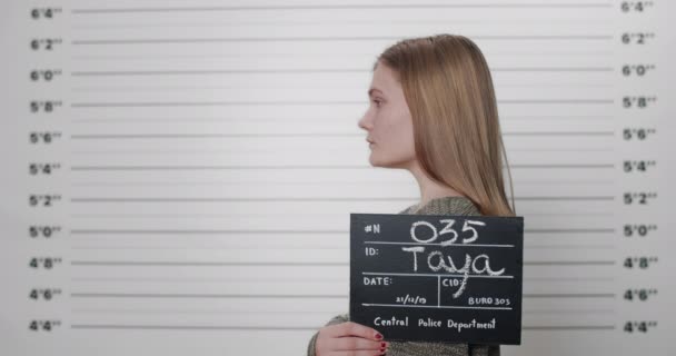 Foto de perfil lateral de una mujer sosteniendo un letrero mientras era fotografiada frente a la pared métrica de la estación de policía. Mujer joven girando la cabeza y mirando a la cámara. — Vídeos de Stock