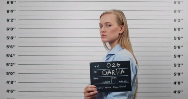 Seitenprofilbild einer attraktiven Frau mit langen Haaren, die den Kopf dreht und in die Kamera blickt. Kriminelle Millennials mit Schild für Foto vor Polizeiwand. — Stockvideo