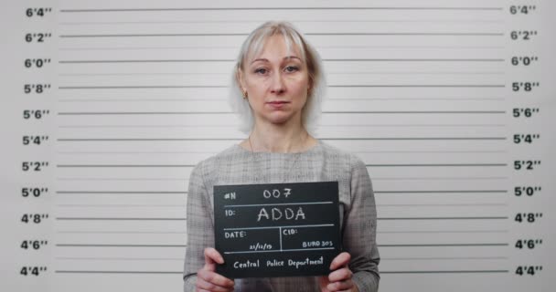 Portrait of blond hair woman holding sign for photo in police department. Mugshot of female criminal in 40s posing, raising head and looking to camera.Concept of crime. — Stock Video