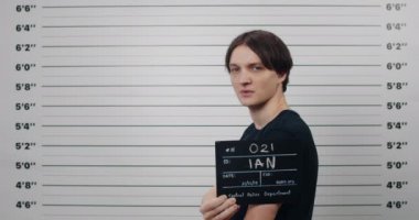 Siyah saçlı, başını çevirip kameraya bakan bir erkeğin yan profili. Polis metrik sıralama duvarının önünde dururken elinde fotoğraf tabelası tutan suçlu genç adam..