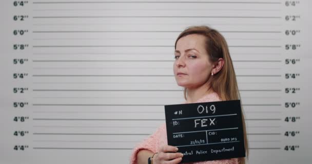 Side profile mugshot of arrested young woman with long hair turning head and looking to camera. Crop view of female criminal in 43s holding sign for photo in police department. — Stock Video