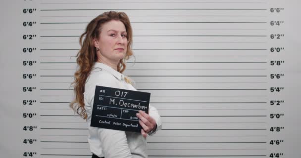 Mugshot of arrested woman with long wavy hair turning head and looking to camera while standing aside. Female criminal in middle 40s holding sign for photo in police department. — Stock Video