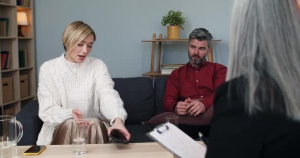 Suspicious woman holding husband phone while he trying taking back smartphone at therapy session.Crop view of married couple having conversation with female family counselor. — Wideo stockowe