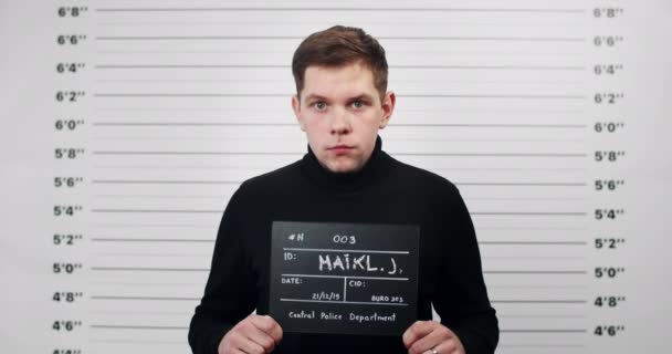 Portrait of guy in black turtleneck holding sign for photo in police department. Crop view of millennial man posing, raising head and looking to camera. Concept of crime. — Stock video