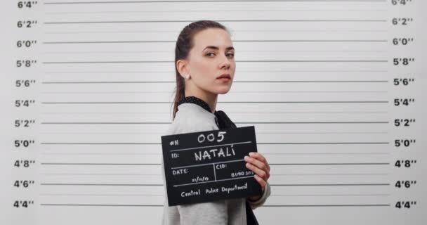 Mugshot of arrested young woman turning head and looking to camera while standing aside. Crop view of pretty female criminal with ponytail holding sign for photo in police department. — Vídeo de Stock