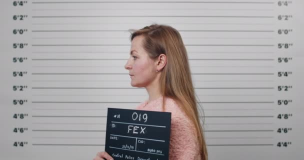 Mugshot of arrested young woman with long hair turning aside and looking to camera while standing near metrical lineup wall . Female criminal in 30s holding sign for photo in police station. — Stockvideo