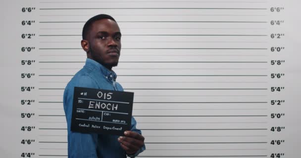 Een politiefoto van een Afrikaanse crimineel die op het politiebureau wordt gefotografeerd. Crop view van man in de jaren '30 draaien hoofd en op zoek naar camera terwijl opzij te staan en houd teken. — Stockvideo