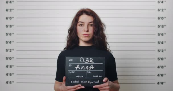 警察署で撮影された巻き毛の逮捕された百万人の女性のムグショット。若い女性犯罪者は頭を上げ、メートルラインの壁の前に立っている間、カメラを見て. — ストック動画