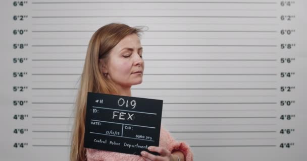 Side profile mugshot of female person with long hair turning head and looking to camera. Criminal woman holding sign for photo while standing in front of police metric lineup wall. — Stock Video