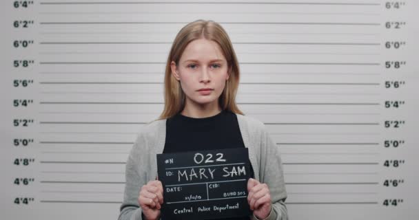 Mugshot of attractive arrested woman holding sign for photo in police department. Crop view of young female criminal posing, raising head and looking to camera in front of metric lineup wall. — Stock Video