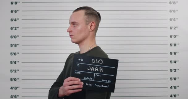 Mugshot av killen i 30-årsåldern vänder sig åt sidorna medan du håller skylt och poserar för foto. Beskär syn på kriminella manliga person med hästsvans tittar till kameran i polisen. Begreppet brott. — Stockvideo