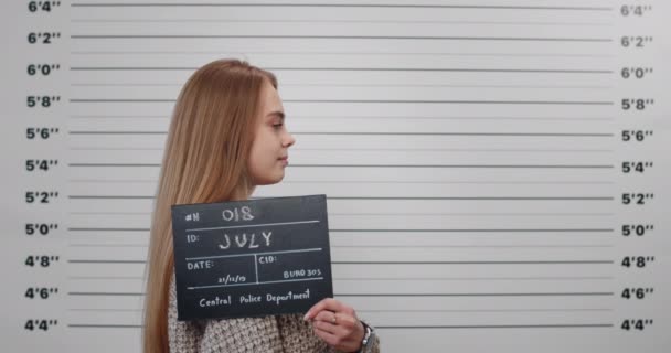 Zdjęcie przestępczej młodej kobiety z długimi blond włosami odwracającymi głowę i patrzącej w kamerę stojąc z boku. Ładna kobieta Peson trzyma znak dla zdjęcia w policji. — Wideo stockowe