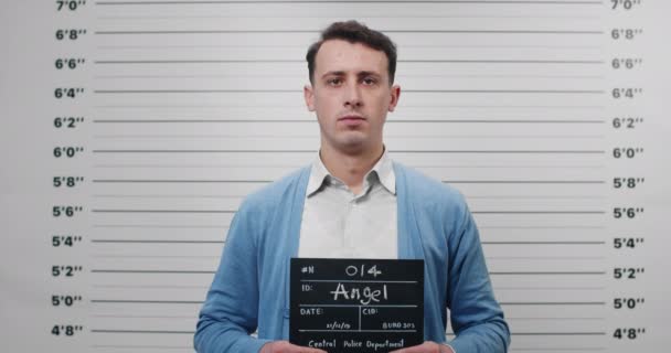 Mugshot de tipo en camisa un cárdigan girando a los lados mientras sostiene el cartel y posando para la foto. Vista de la cosecha de hombre criminal con el pelo corto ondulado mirando a la cámara en el departamento de policía. — Vídeo de stock