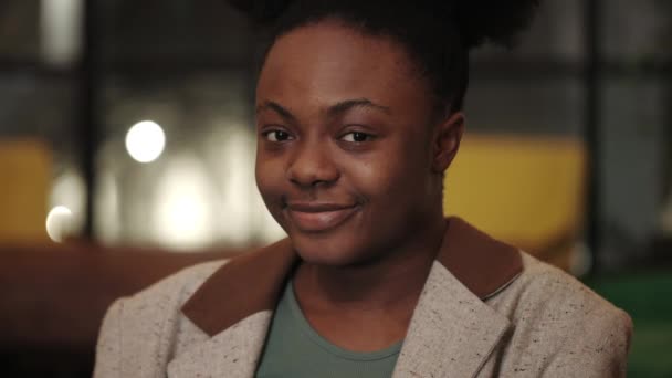 Vista de la cosecha de la joven mujer afro-americana alegre en chaqueta girando la cabeza y sonriendo mientras mira a la cámara. Retrato de una persona positiva posando. Fondo borroso. — Vídeo de stock