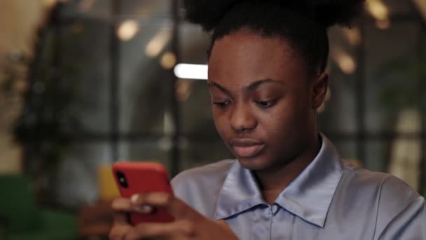 Vista de la cosecha de la mujer afroamericana en blusa con teléfono inteligente mientras está sentada en la silla. Persona bastante femenina navegando por Internet y escribiendo en la cafetería mientras pasa tiempo libre. — Vídeo de stock