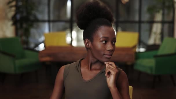 Vista de la cosecha de la joven mujer afro-americana alegre apoyando y girando la cabeza mientras mira a la cámara. Retrato de una hermosa mujer posando y sonriendo en la cafetería. — Vídeo de stock
