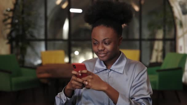 Vista de la cosecha de la mujer afroamericana en blusa con teléfono inteligente mientras está sentada en la silla. Atractiva persona femenina sonriendo y navegando por Internet en la cafetería mientras pasa tiempo. — Vídeos de Stock
