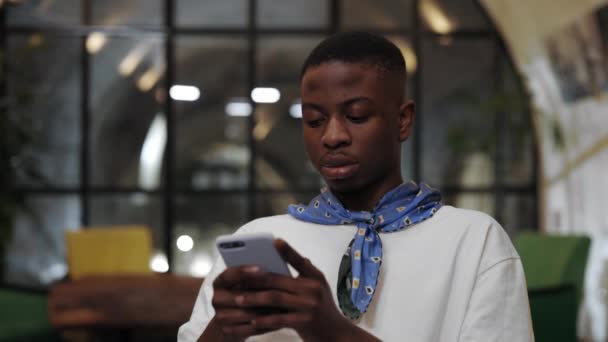 Vista de la cosecha de un joven afroamericano usando un teléfono inteligente mientras está sentado en la cafetería. Hombre guapo con pañuelo escribiendo mensaje mientras charlaba en las redes sociales. Concepto de ocio. — Vídeos de Stock