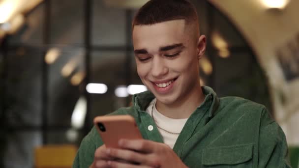 Vista de perto do jovem com corte de cabelo elegante usando smartphone enquanto sentado na cadeira. Bonito homem em roupas casuais rindo enquanto rola notícias de mídia social no café. — Vídeo de Stock