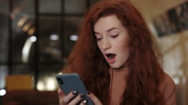 Bin yıllık kızıl kıvırcık saçlı, hayrete düşmüş ve kafede akıllı telefon kullanırken 