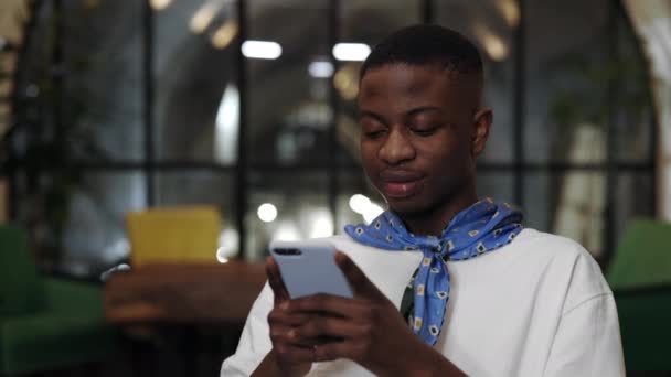 Recorte de vista de millennial afro americano chico usando teléfono inteligente y riendo mientras está sentado en la cafetería. Lindo hombre con pañuelo escribiendo mensaje mientras charlas en las redes sociales. Concepto de ocio. — Vídeo de stock