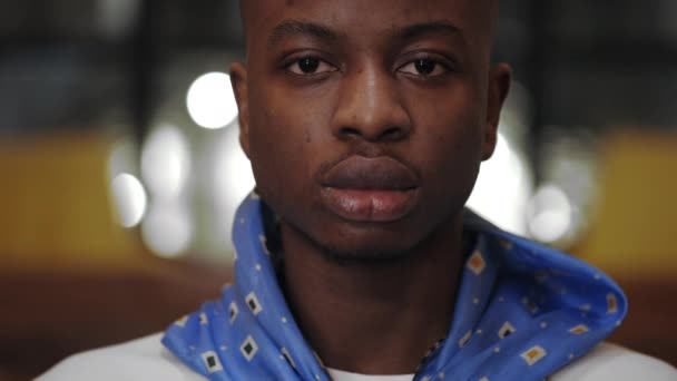 Vue rapprochée du jeune homme afro-américain sérieux regardant vers la caméra. Portrait d'un bel homme avec un foulard posant dans un café. Fond flou. — Video