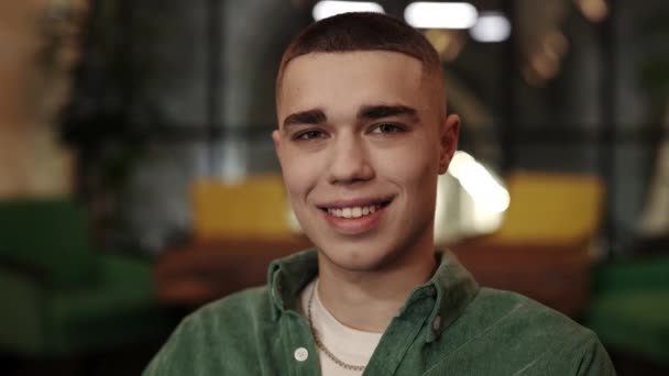 Vista de la cosecha de hombre joven con corte de pelo elegante sonriendo mientras mira a la cámara. Retrato de chico alegre guapo posando mientras está sentado en la café.Concepto de emociones positivas. Fondo borroso. — Vídeos de Stock