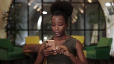 Cafe 'de akıllı telefon kullanan Afro-Amerikan bir kadının görüntüsüne yakından bakın. Sosyal medyaya kayan genç kadın haberleri ve duygularını gösteriyor.