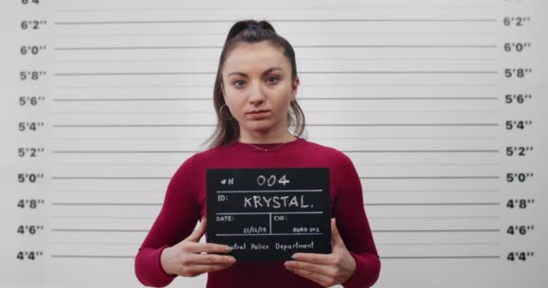 Mugshot of millennial woman with ponytail turning to sides while posing for photo. Portrait of female criminal looking to camera and holding sign in police department.Concept of crime. — Stock Video