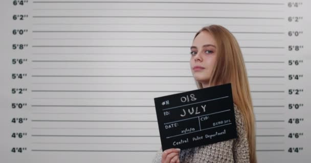 Mugshot einer kriminellen Millennial-Frau mit langen blonden Haaren, die den Kopf dreht und im Stehen in die Kamera schaut. Attraktive Pesonin hält Schild für Foto bei der Polizei hoch. — Stockvideo