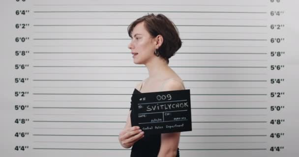 Mugshot de una joven arrestada con corte de pelo corto girando la cabeza y mirando a la cámara mientras gira a diferentes lados. Morena mujer criminal sosteniendo cartel para la foto en el departamento de policía. — Vídeos de Stock