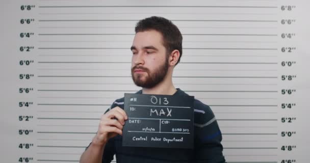 Mugshot de la personne mâle millénaire se tournant vers les côtés tout en tenant signe et posant pour la photo. Vue de la récolte de l'homme arrêté avec barbe et moustaches regardant à la caméra dans le département de police. — Video