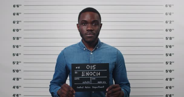 Portret afro amerykańskiego mężczyzny trzymającego tabliczkę ze zdjęciem na posterunku policji. Widok na młodego człowieka pozującego, podnoszącego głowę i patrzącego w kamerę. Pojęcie zdjęcia policyjnego i przestępstwa. — Wideo stockowe