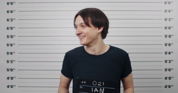 Retrato de un joven detenido sosteniendo un letrero para la foto en el departamento de policía. Mugshot de chico hablando y amenazando mientras mira a la cámara frente a la pared métrica de alineación. — Vídeos de Stock