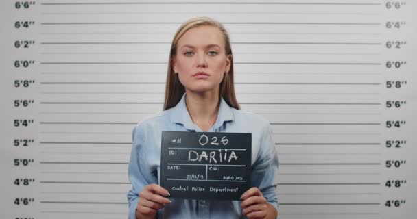 Een politiefoto van een mooie vrouw met een bord voor foto op het politiebureau. Crop view van jonge vrouwelijke crimineel raisen hoofd en op zoek naar camera terwijl staande voor de metrische line-up muur. — Stockvideo