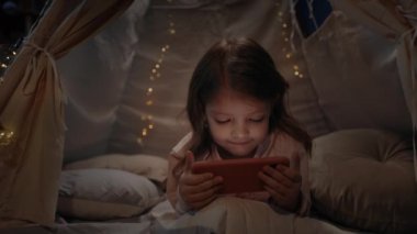 Sevimli küçük kız akşamları evde ışık saçan dekoratif bir kulübede vakit geçiriyor. Çocuk akıllı telefondan çizgi film izlerken yerde yatıyor. Dikkatsiz çocukluk kavramı.