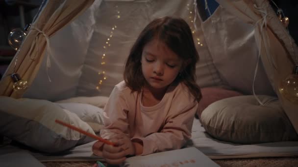 Comely menina desenho com lápis de cor, enquanto deitado no chão em bela tenda decorativa em casa. Criança bonito passar o tempo livre à noite. Conceito de infância despreocupada. — Vídeo de Stock