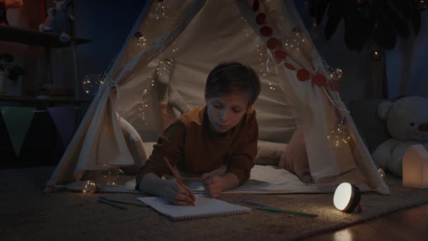 Positive boy drawing with colored pencils on white paper shit while lying on floor in decorative tent at home. Male teen spending free time in evening. Concept of creativeness. — Stock Video